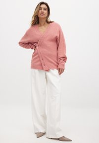 Cárdigan de punto rosa con escote en V profundo, cierre frontal de botones y puños acanalados, combinado con pantalones blancos de pierna ancha.
