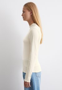 Rippentop mit langen Ärmeln in hellem Cremeton, mit einem Stehkragen und gerafftem Saum; kombiniert mit hellblauen Jeans.