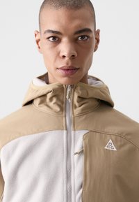 Beige og lys grå jakke med hette, med glidelås-lukkning og trekantet logo. Myk tekstur med en jevn finish og minimalistisk design.