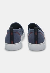 Pantofi slip-on din pânză albastră, cu talpă de cauciuc albă și cusături roșii în contrast. Prezintă un tab de prindere negru cu textul "RED LABEL" pe spate.