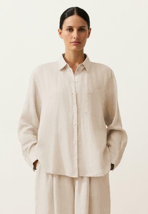 Femeie cu păr închis la culoare, purtând o cămașă largă bej cu nasturi și pantaloni asortați, pe un fundal simplu, deschis la culoare.