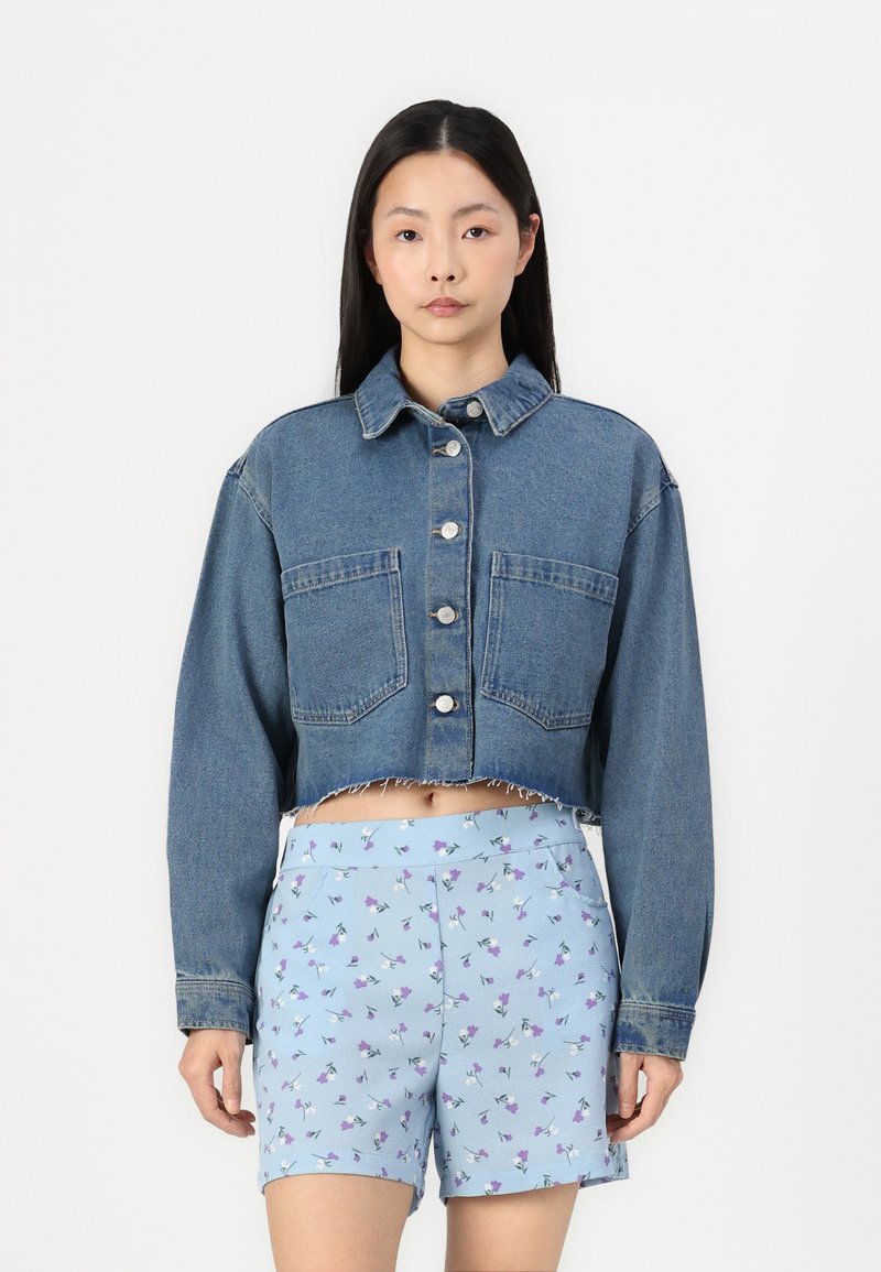 Κατακόκκινη τζιν ζακέτα με μεγάλες τσέπες, κλείσιμο με ασημένια κουμπιά και φθαρμένο στρίφωμα. Συνδυάζεται με ανοιχτό μπλε σορτσάκι με λουλουδάτο σχέδιο.