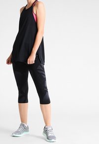 Svart träningströja i lös passform; tillsammans med svarta capribyxor med geometriskt mönster. Grå sneakers med gröna detaljer.