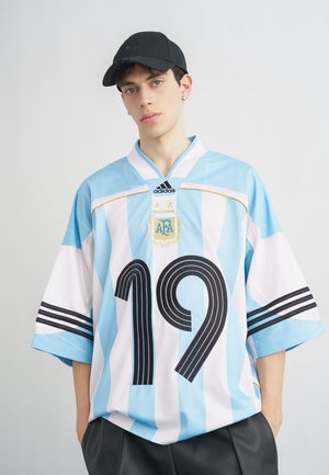 Fiatal férfi fekete sapkában, túlságosan nagy, világoskék-fehér csíkos Argentína focimezben, rajta a 19-es szám és az Adidas logó.