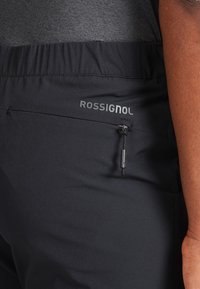 Pantaloni tecnici neri con vita elastica, tasca con zip e logo "ROSSIGNOL" stampato. Materiale liscio con finitura opaca.
