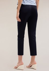 Marineblauwe tapered broek met achterzakken, gemankeerde enkels en een gladde textuur, gecombineerd met lichte wedgesandalen.