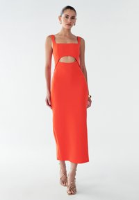 Vrouw in een mouwloze, feloranje midi-jurk met een uitsparing bij de buik, gecombineerd met gouden sandaaltjes en gouden sieraden tegen een witte achtergrond.