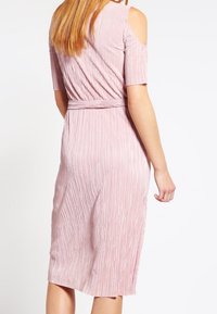 Frau in einem knielangen, hellrosa, plissierten Kleid mit kurzen Kalt-Schulter-Ärmeln, die mit dem Rücken zur Kamera steht.