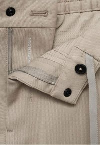 Pantalon en tissu beige avec fermeture éclair, fermeture par bouton, doublure intérieure de poche et texte visible indiquant "NING PERFORM."