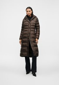 Καφέ φούτερ παλτό με οριζόντια ραφή, μήκους μέχρι το γόνατο, με φερμουάρ και κουμπί, κουκούλα, φορεμένο πάνω από σκούρα τζιν και μποτάκια με τακούνι.