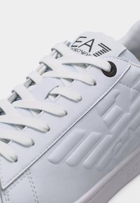Κοντινό πλάνο λευκού αθλητικού παπουτσιού με ανάγλυφο λογότυπο «EA7», λευκά κορδόνια και μαύρες τρύπες κορδονιών σε λείο δερμάτινο πάνω μέρος.