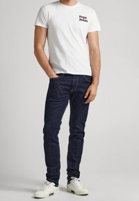 Vit t-shirt med "Pepe Jeans"-logotypen på bröstet, matchad med mörkblå jeans och vita sneakers med gröna detaljer.