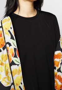 Schwarzes Oberteil mit kurzen Ärmeln, kombiniert mit einer cardigan im Blumenmuster, die gelbe und orangefarbene Blumen auf einem schwarzen Hintergrund zeigt.