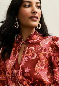 Blouse rouge à motif paisley avec des manches bouffantes, un col à volants et une ouverture en forme de goutte, fabriquée en tissu lisse et brillant. Grandes boucles d'oreilles en or.