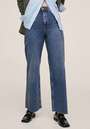 Personne portant un jean bleu large taille haute, des mocassins noirs, une chemise rayée bleu clair partiellement rentrée et une veste verte, debout.