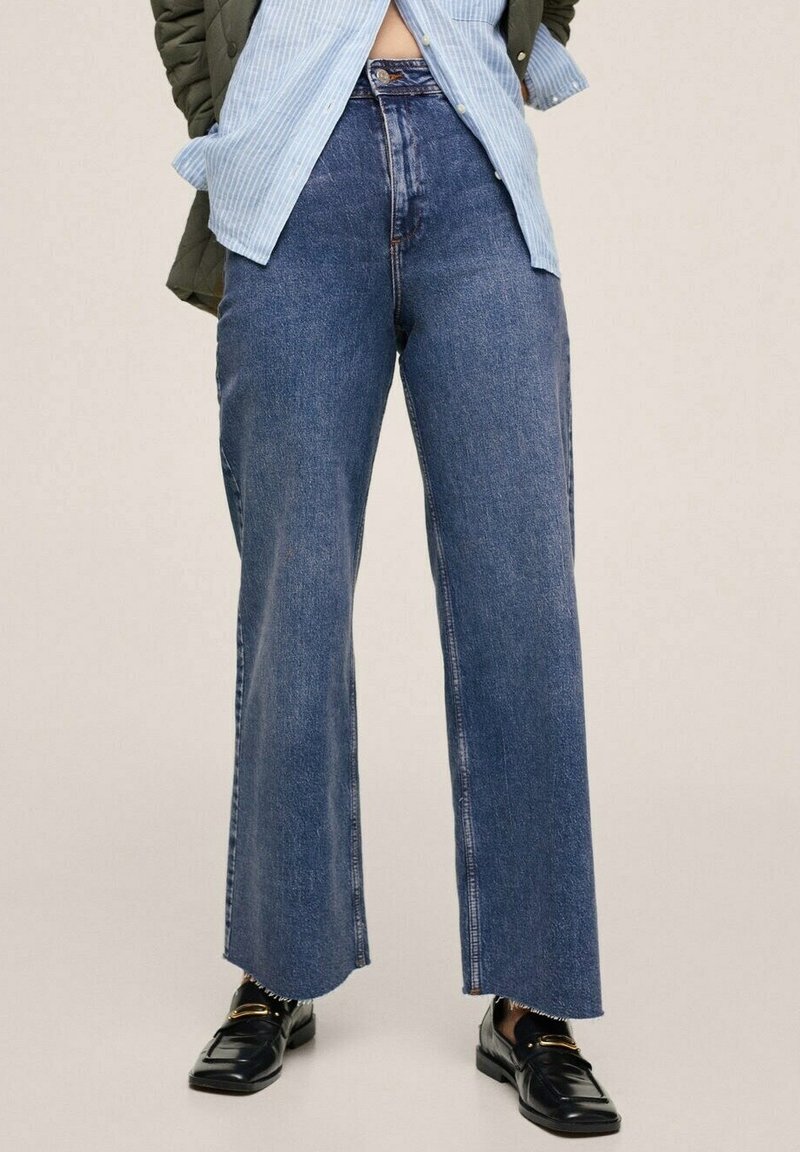 Personne portant un jean bleu large taille haute, des mocassins noirs, une chemise rayée bleu clair partiellement rentrée et une veste verte, debout.