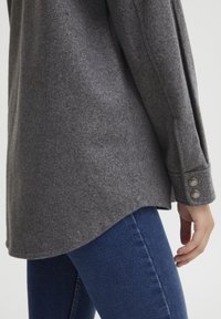 Szary top z mieszanki wełny z zaokrąglonym dołem, długimi rękawami i dwoma guzikami na mankietach, w zestawieniu z niebieskimi dżinsami.