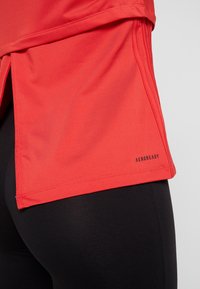Červené sportovní tričko s delším zadním lemem, krátkými rukávy a značkou AeroReady na boku, v kombinaci s černými legínami. Hladká textura.