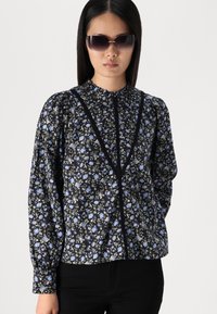 Blusa nera a fiori con fiori blu, maniche lunghe, colletto arricciato e dettagli scuri. Abbinata a pantaloni neri e occhiali da sole.
