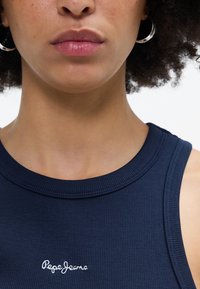 Marineblå ribbestrikket tanktop med rund hals og hvit "Pepe Jeans" logo brodert på brystet. Har en tettsittende design og glatt tekstur.
