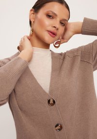 Jasnobrązowy kardigan z głębokim dekoltem w serek, prążkowanymi rękawami i trzema zapięciami na guziki. Miękka faktura, luźny krój i subtelne detale na ramionach.