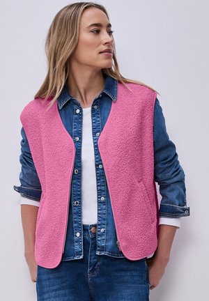 Donna che indossa un gilet senza maniche in pile rosa acceso sopra una giacca di jeans blu con maniche arrotolate, che guarda verso sinistra.