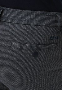 Close-up of a dark gray textured fabric pocket with a single black button and subtle brand logo on tailored trousers.
