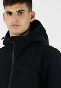 Giacca nera impermeabile con colletto alto, cappuccio regolabile e zip. Presenta un logo discreto in stampa scura sul davanti.