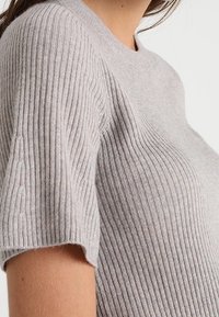 Světle šedý ribbed úpletový top s krátkými rukávy a kulatým výstřihem. Textura má vertikální žebrování, což poskytuje subtílní vzorový detail.