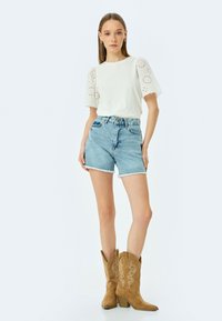 Hvid skjorte med øjeborede pufærmer, blå denim shorts med frynset kant og brune cowboystøvler. Simpelt design med mærkbar detaljering.