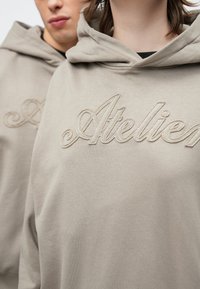 Bézs színű pamut kapucnis pulóver, amelyen elöl nagy, texturált "Atelier" hímzés található. Kapucnis kialakítás lágy, sima anyaggal.