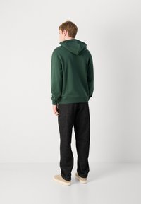 Felpa verde con tasca a marsupio, polsini e orlo a coste, abbinata a pantaloni grigi scuri e sneakers beige. Sfondo semplice.
