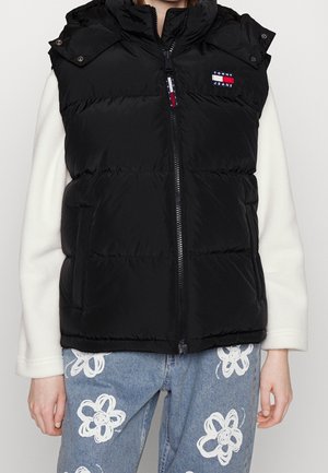 Osoba ubrana w czarną pikowaną kamizelkę z logo Tommy Jeans na białej, długorękawowej bluzce oraz niebieskie jeansy z białymi kwiatowymi wzorami.