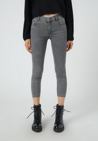 Jeans de mezclilla gris con un diseño ajustado y longitud recortada, que presentan bolsillos frontales y un dobladillo sin acabar. Combinados con botas negras con cordones.