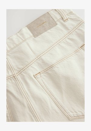 Beige pamut nadrág vékony függőleges csíkokkal, bőr folttal, amin a "DENIM SUPPLY" felirat szerepel, és ellentétes színű varrással a zsebeken.