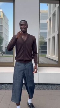 Mladý muž stojí vonku, oblečený v hnedom kardigane, širokých skrátených džínsoch, bielych ponožkách a čiernych mokasínach, s modernými budovami v pozadí.