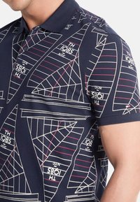 Camisa polo navy con un patrón geométrico de veleros en blanco y rojo, con un cuello clásico y mangas cortas. Tejido de algodón suave.