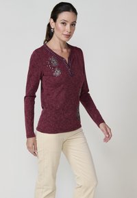 Burgundijas garo piedurkņu tops ar ziedu izšuvumiem, V veida kakla izgriezumu un dekoratīvām pogām. Apvienots ar krēma krāsas korduroja biksēm. Mīksta auduma tekstūra.