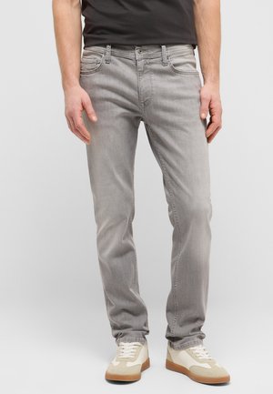 Mann trägt hellgraue Slim-Fit-Jeans, weiße und beige Sneakers, steht vor einem schlichten hellen Hintergrund.