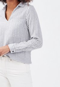 Chemise à rayures grise et blanche. Dispose d'un col en V, de manches longues, d'une poche et de subtils accents métalliques. Tissu doux et léger.