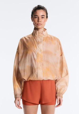 Nő laza, őszibarack tónusú magas nyakú dzsekit visel hosszú ujjakkal és rozsda színű rövidnadrággal, sima fehér háttér előtt.