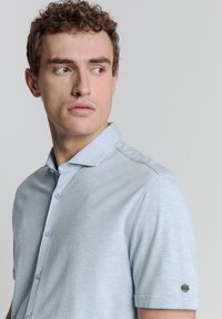 Hellesblaues, kurzärmeliges Polo-Shirt mit einem strukturierten Kragen, ausgestattet mit einer Knopfleiste mit zwei Knöpfen und einem Logo-Akzent am linken Ärmel.