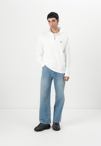 Vitt zip-neck sweatshirts i tyg, parat med ljusblå vida jeans och svarta sneakers. Inkluderar en grön logotyp på bröstet.