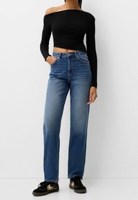 Černý top s odhalenými rameny a dlouhými rukávy, který je zkombinován s modrými širokými jeansy s vysokým pasem. Tenisky mají černobílý design.