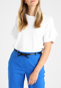 Top blanco de manga corta con bordes con volantes, combinado con pantalones azul brillante, presentando un cinturón negro con un detalle de lazo.