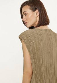 Abito khaki plissettato con maniche a cappuccio, caratterizzato da una texture verticale e una chiusura posteriore discreta. Tessuto morbido e design fluido.