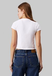 Biely bavlnený crop top s krátkymi rukávmi, nosený s vysokými modrými džínsami s koženou nášivkou a zadnými vreckami.