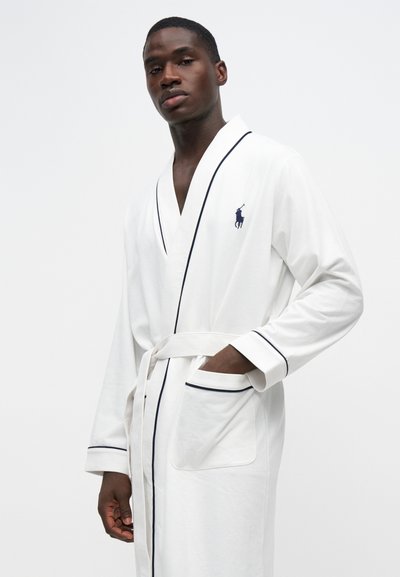 Homme portant une robe blanche avec des bordures noires et un logo sur la poitrine, debout avec une main dans la poche devant un fond uni.