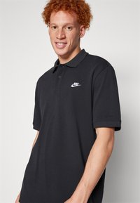 Fekete polo póló, struktúrált anyagból, három gombos kivágással és egy kis fehér Nike logóval a bal mellrészen.