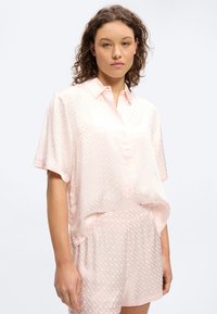 Mladá žena s kudrnatými vlasy, která má na sobě světle růžovou saténovou košili s krátkými rukávy na knoflíky a odpovídající šortky s jemným geometrickým vzorem.